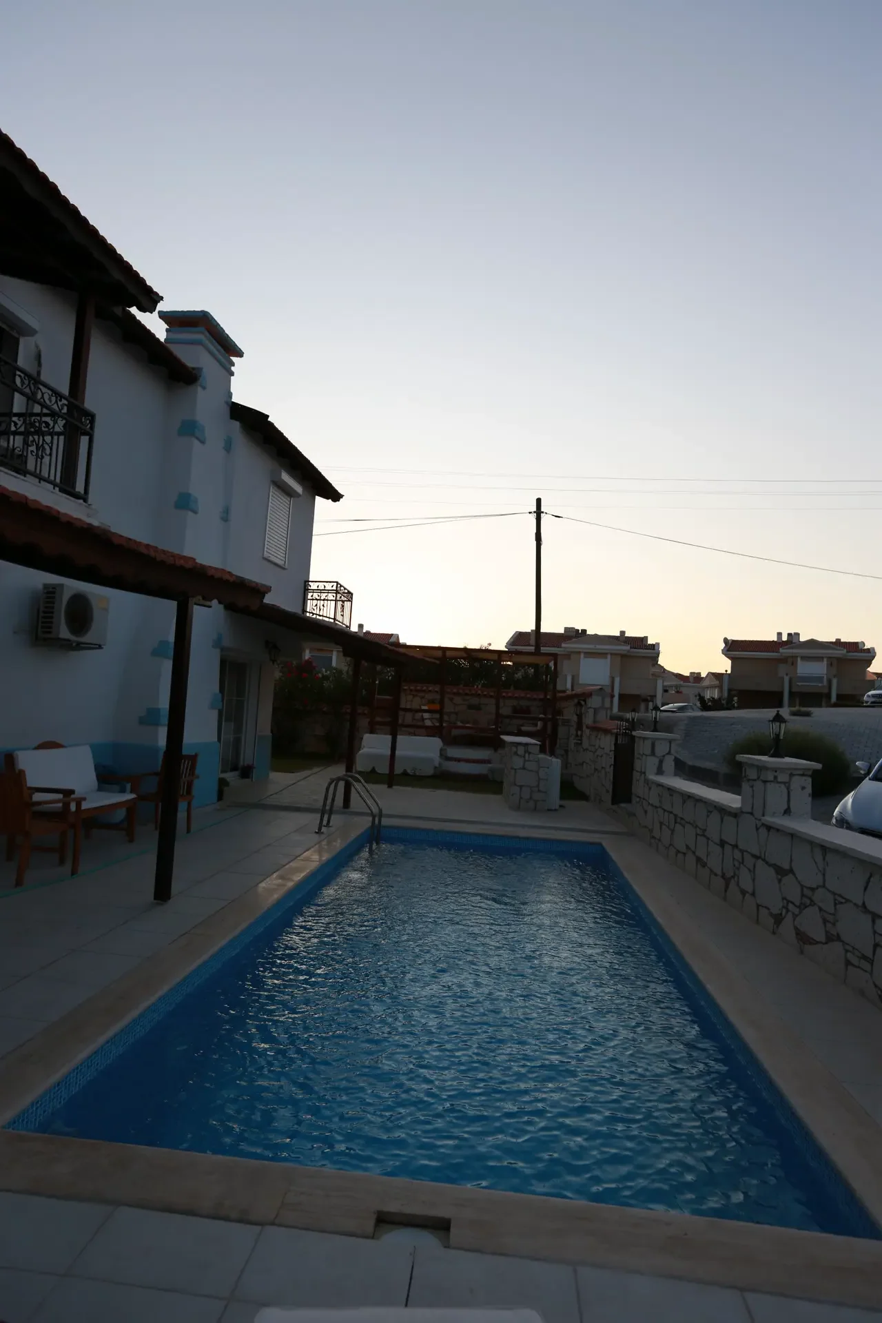 Köşeli formda, koyu granit kaplamalı bir Fiplus havuzu; paslanmaz şelale ağzı, ahşap deck ve bahçe düzenlemesiyle